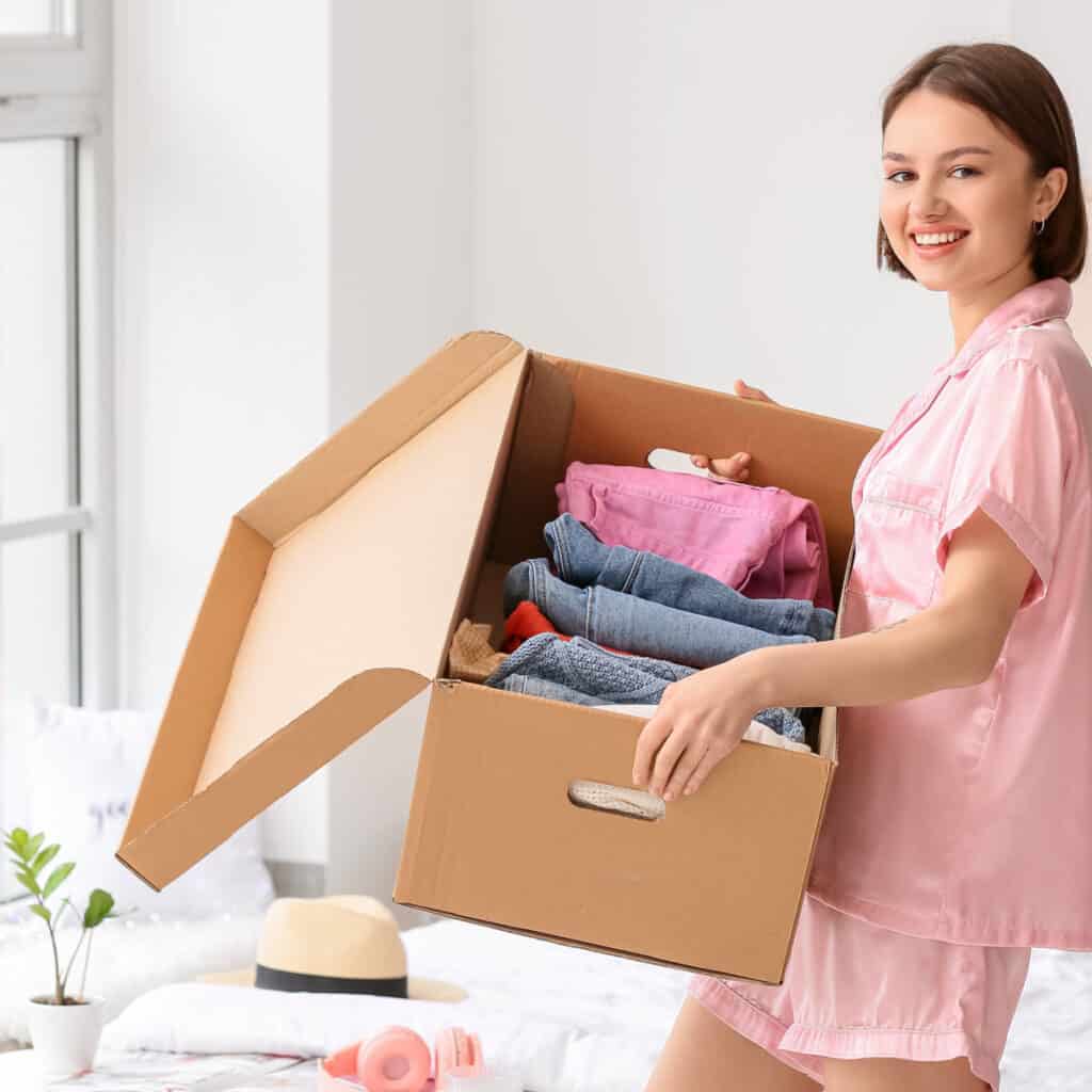 smiling woman holding large open box with neatly folded clothing in bright neat room.