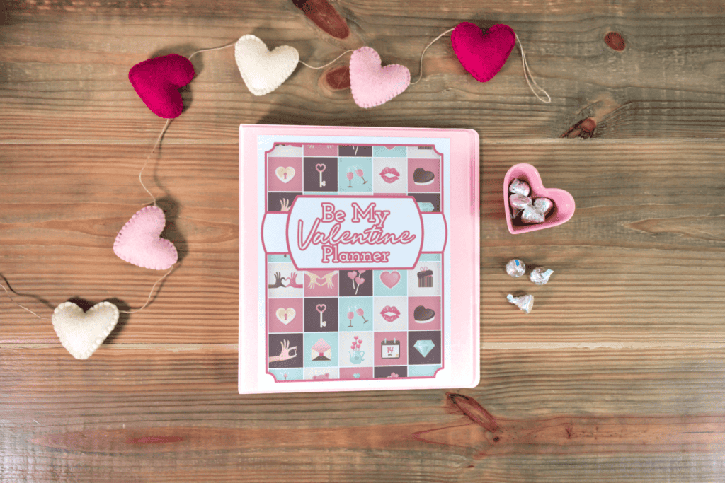 A &ldquo;Be My Valentine Planner&rdquo; notebook on a wooden table, surrounded by heart-shaped decorations and a heart-shaped dish with silver-wrapped chocolates.