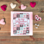 A &ldquo;Be My Valentine Planner&rdquo; notebook on a wooden table, surrounded by heart-shaped decorations and a heart-shaped dish with silver-wrapped chocolates.
