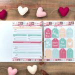 An open binder on a wooden table displays Valentine-themed pages, surrounded by a string of plush pink, red, and white hearts.