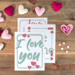 Three &ldquo;I love you&rdquo; cards on a wooden table with heart-shaped decorations, a pink heart dish containing candies, and a garland of fabric hearts.